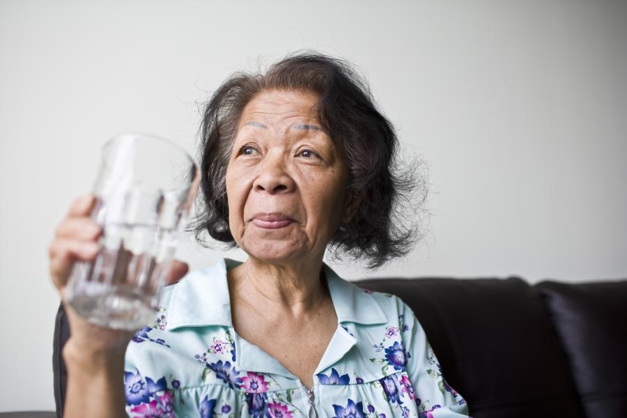 An elderly Asian woman drinks a glass of water while sitting on a couch