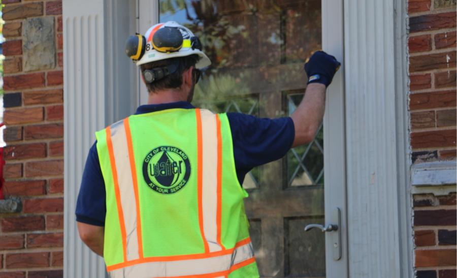 A man wearing a hard hat and yellow high-visibility vest with a Cleveland Water logo on the back knocks on the front door of a house.