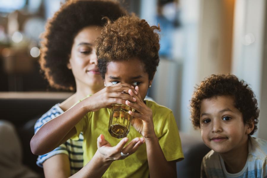 A mom sits on a couch with her two boys. One is sitting on her lap drinking a glass of water.