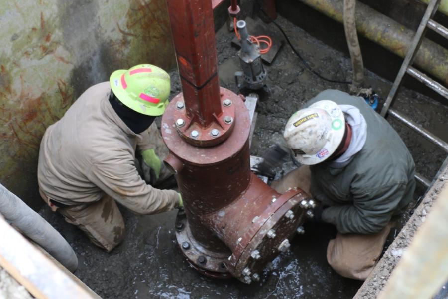 Two men wearing hard hats in a trench work on repairing a water main break